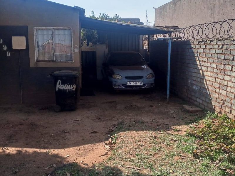 Rear exterior view showing a basic carport structure, brick boundary wall with security razor wire, and an unpaved yard area.