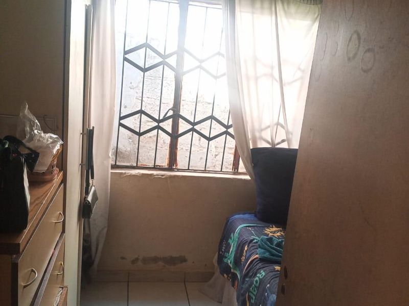 A compact bedroom featuring tiled flooring, a built-in cupboard, and a window fitted with a security gate, requiring cosmetic updates and maintenance.