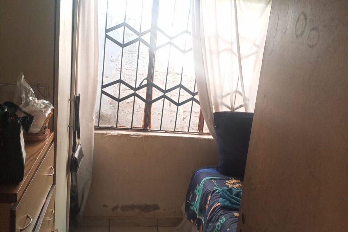 A compact bedroom featuring tiled flooring, a built-in cupboard, and a window fitted with a security gate, requiring cosmetic updates and maintenance.