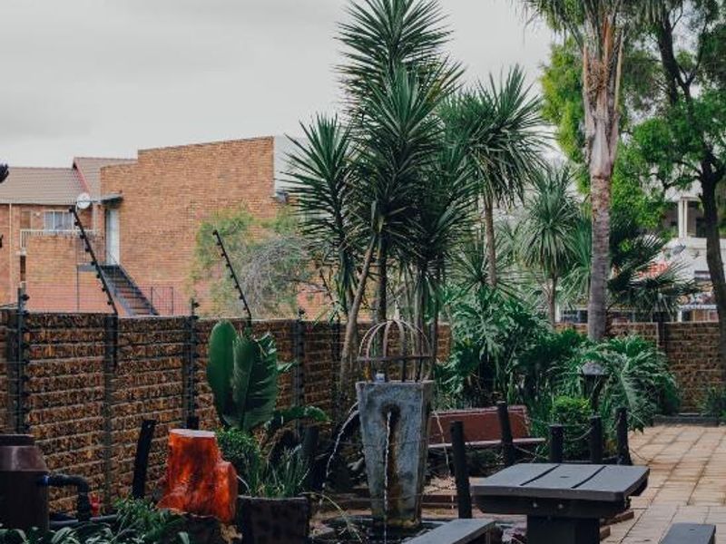 A landscaped garden area featuring a paved patio, established palm trees, a stone water feature, and secure brick boundary walls with electric fencing.
