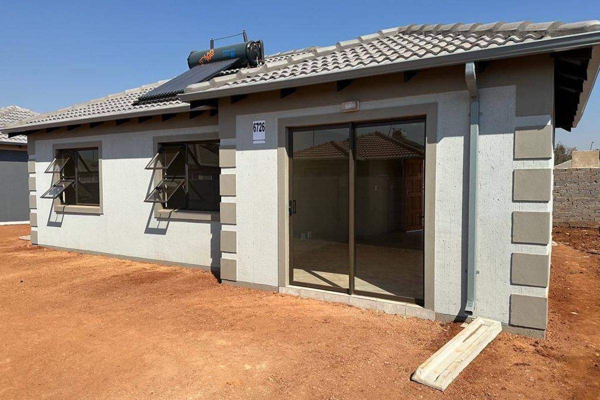 Rear view of a modern single-story house featuring a tiled roof, solar water heater, sliding glass door, and casement windows with unfinished surrounding grounds.