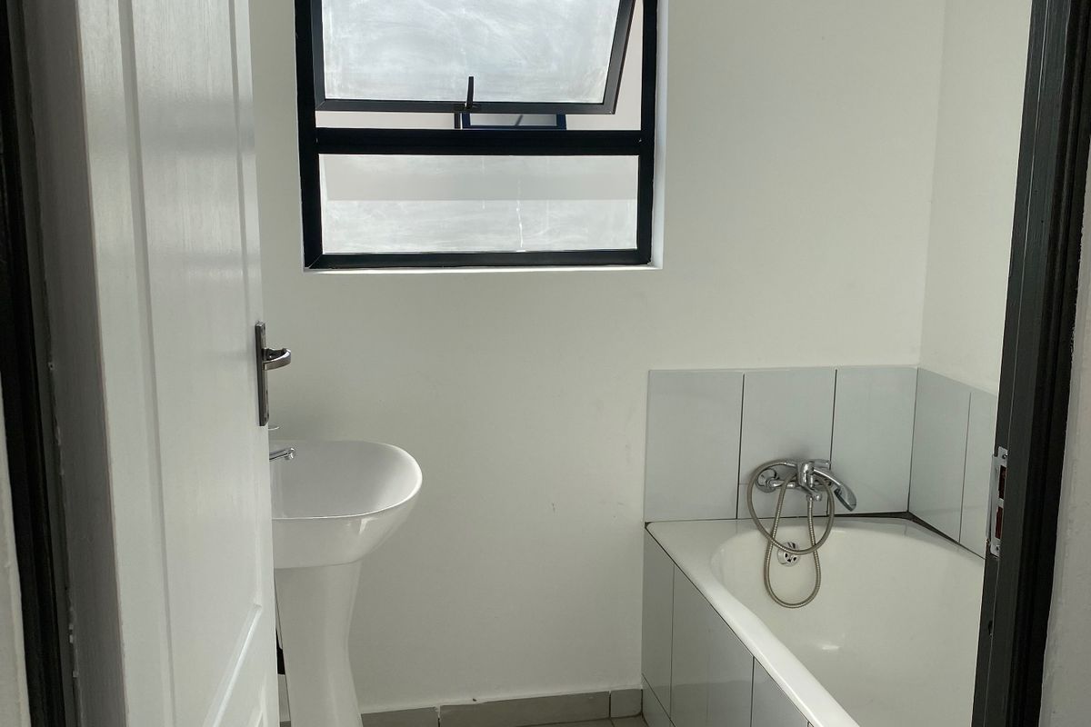 A clean, functional bathroom featuring a white pedestal basin, a tiled bathtub, and neutral floor tiles with a top-hung window for natural light.