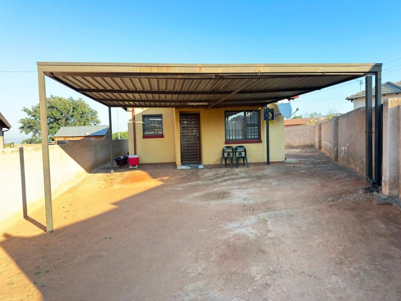 Front exterior view of the property featuring a large metal carport, security gate, and perimeter walling on an unpaved driveway.
