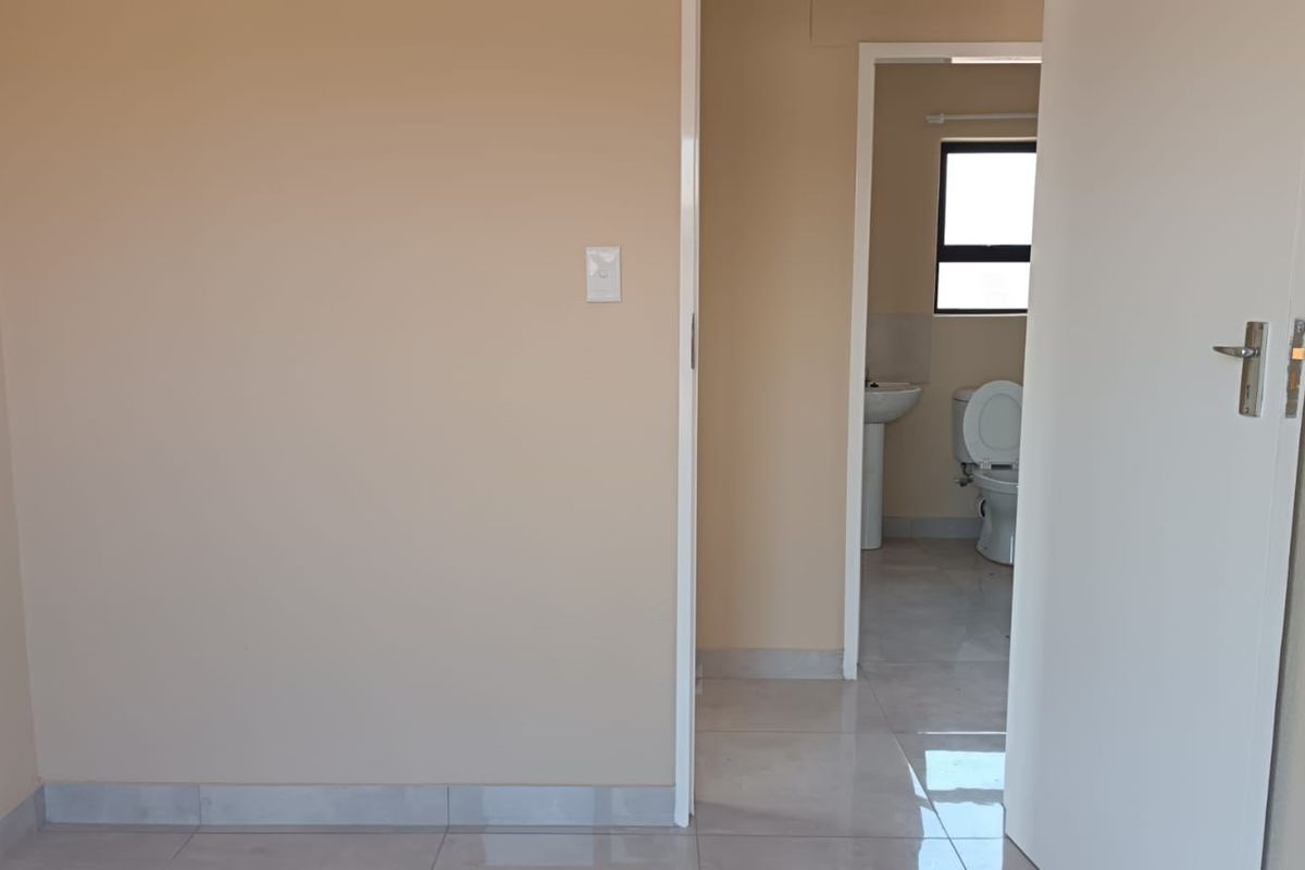 A clean, empty bedroom featuring neutral wall tones and tiled flooring, with an adjacent en-suite bathroom visible through the doorway.