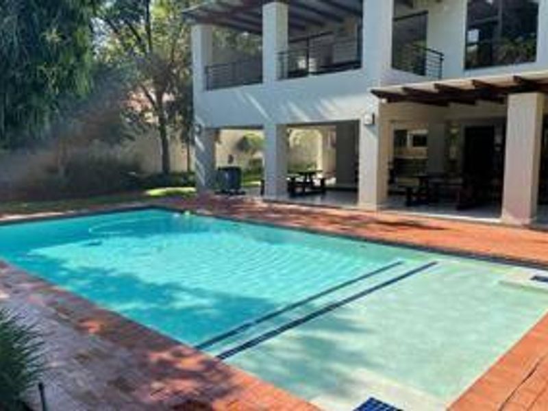 A well-maintained swimming pool area featuring brick paving, a spacious covered patio, and an upper-level balcony overlooking the outdoor space.