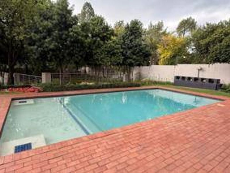 A well-maintained swimming pool area featuring brick paving, surrounding mature trees, and a solid boundary wall in a private garden setting.