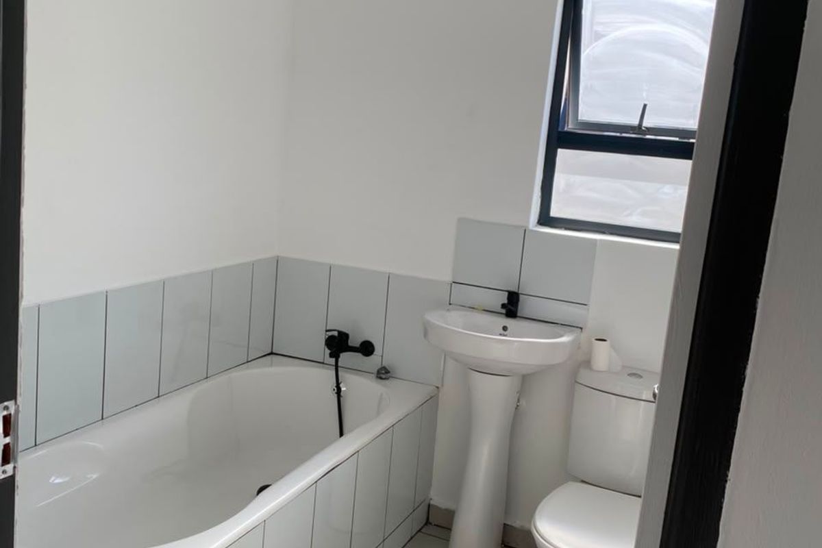 A clean, functional bathroom featuring a white bathtub, pedestal basin, toilet, and neutral floor tiles with a modern black-framed window.