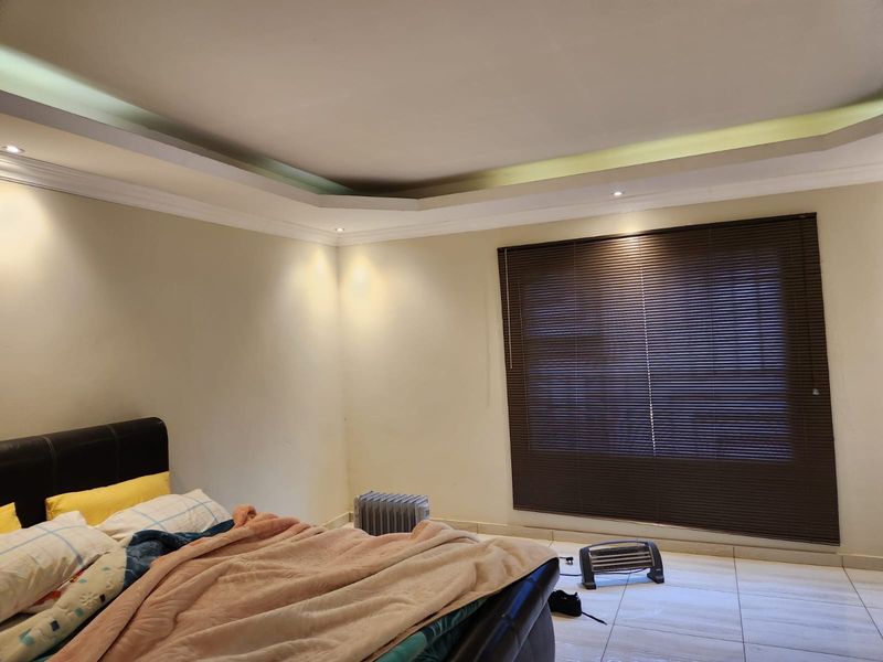 Spacious bedroom featuring tiled flooring, a recessed ceiling with integrated cove lighting, and dark venetian blinds.