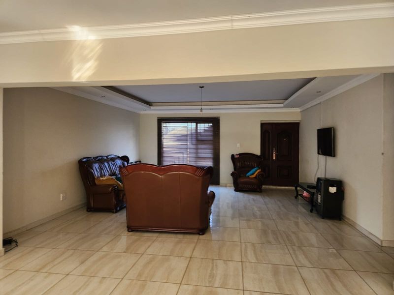 Spacious living area featuring large format floor tiles, recessed ceiling details, and a solid wood entrance door with natural light through venetian blinds.
