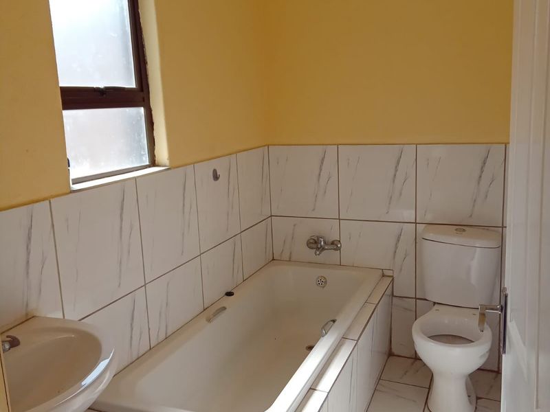 A functional bathroom featuring a built-in bathtub, pedestal basin, and toilet with neutral tiled finishes and natural light from a side window.