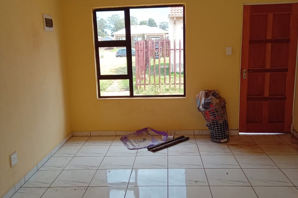 An empty living room featuring light-colored tiled flooring, yellow painted walls, and a large aluminium-framed window providing natural light.
