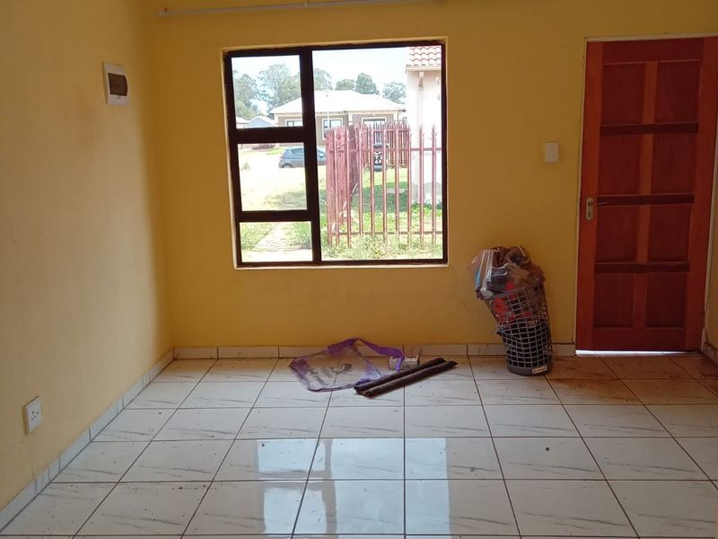An empty living room featuring light-colored tiled flooring, yellow painted walls, and a large aluminium-framed window providing natural light.
