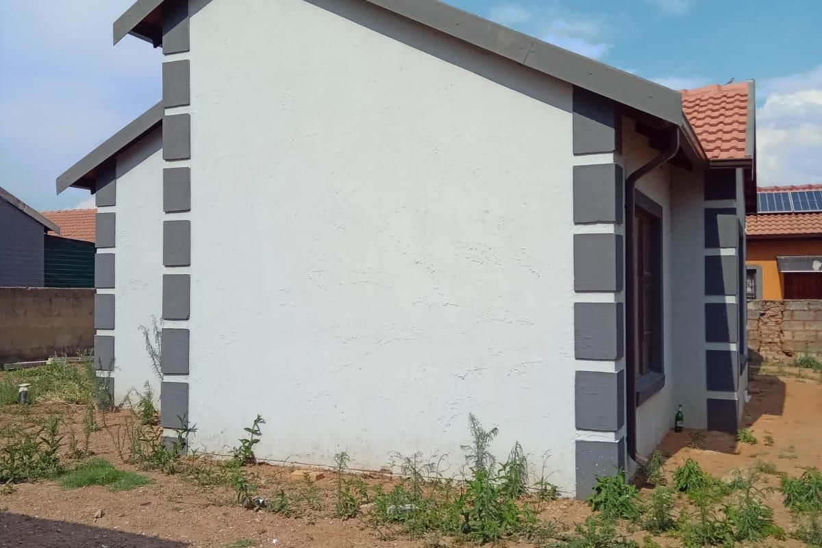 Side and rear view of a residential house featuring a tiled roof, painted exterior walls with decorative corner detailing, and an unlandscaped yard.