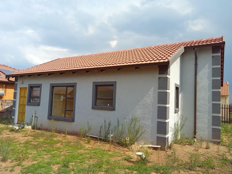 Rear view of a single-story residential house featuring terracotta roof tiles, grey textured plaster walls with contrasting corner detailing, and aluminium window frames.