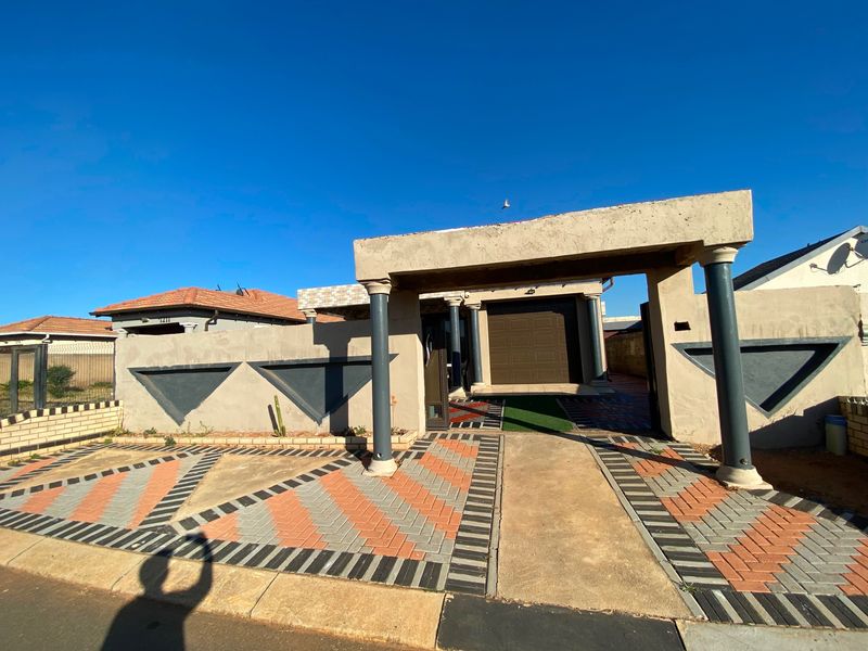 Front elevation of the property featuring a paved driveway, covered entrance with decorative columns, and a secure boundary wall with a garage.