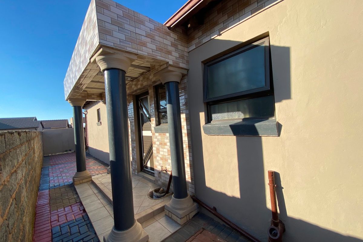 A rear exterior view featuring a covered patio area with decorative columns, tiled facade detailing, and paved ground surfaces.