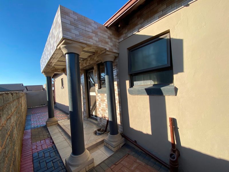 A rear exterior view featuring a covered patio area with decorative columns, tiled facade detailing, and paved ground surfaces.
