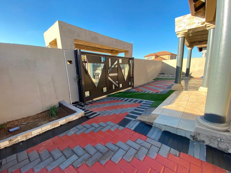 Front entrance area featuring a modern sliding gate, paved driveway with artificial grass accents, and a covered patio with tiled flooring and concrete columns.