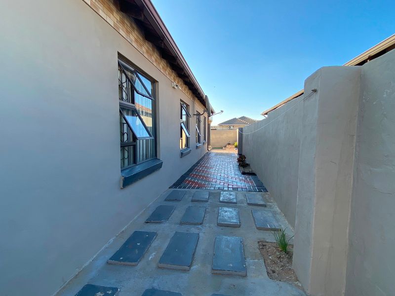 A side exterior walkway featuring paved pathways, boundary walls, and casement windows with security bars along the side of the house.