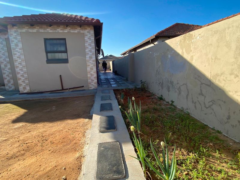 A side exterior view of the property featuring a paved walkway, boundary wall, and unfinished garden area.