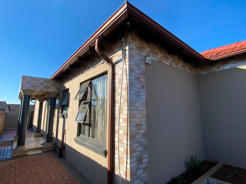 A well-maintained rear exterior view featuring brick facade detailing, a tiled roof, paved patio area, and structural columns.