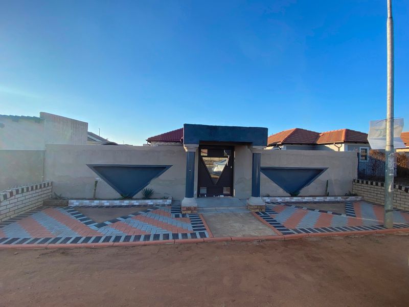 Front view of the property featuring a decorative boundary wall, paved driveway, and a gated entrance with structural pillars.