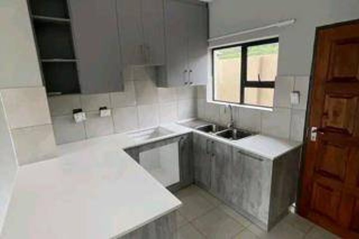 A modern, empty kitchen featuring grey cabinetry, stone countertops, a double sink, and a tiled backsplash with a window for natural light.