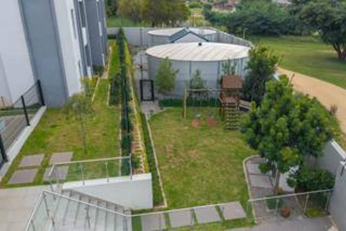 Aerial view of the rear garden area featuring landscaped lawn, paved pathways, glass balustrades, and large water storage tanks at the rear boundary.