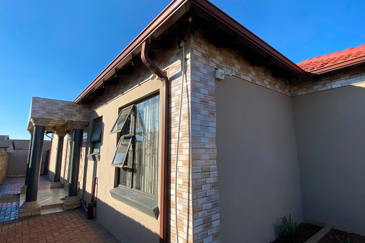 A well-maintained rear exterior view of the house featuring brick facade detailing, a tiled roof, and a paved patio area.
