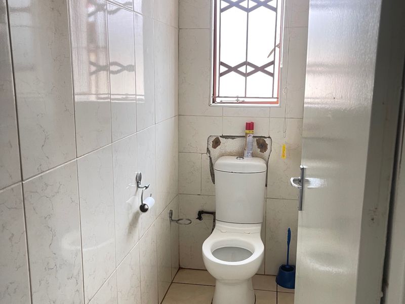 A small, tiled bathroom featuring a standard toilet and a window with security bars, currently requiring minor repairs to wall finishes and floor tiling.