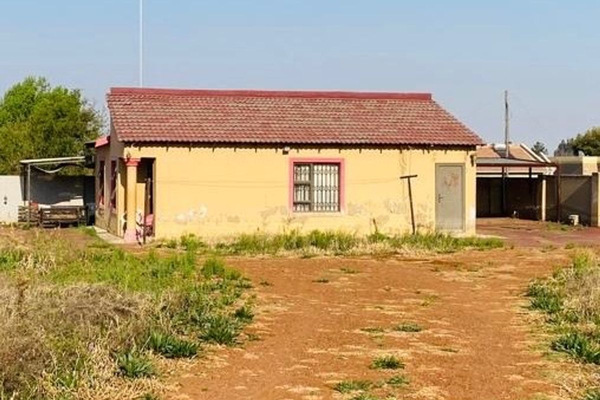 Rear view of a single-story house featuring a tiled roof, security bars on windows, and an attached carport structure on a large plot.
