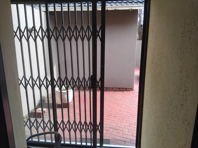A small, paved outdoor patio area viewed through a security gate, featuring plastered walls and a simple exterior layout.