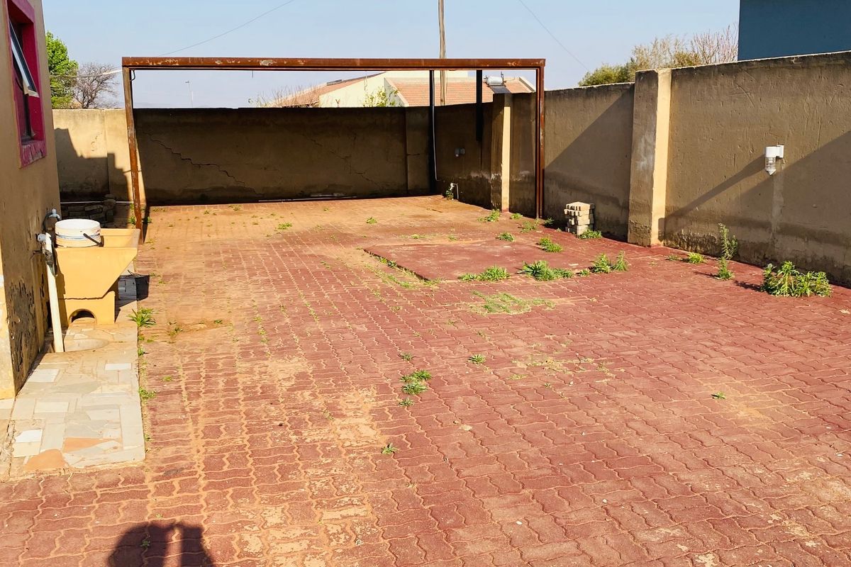 Rear exterior area featuring a paved driveway, metal carport structure, boundary walls, and a utility sink, requiring maintenance and landscaping.