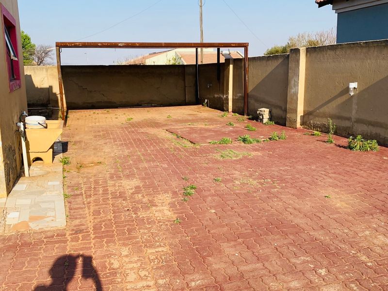 Rear exterior area featuring a paved driveway, metal carport structure, boundary walls, and a utility sink, requiring maintenance and landscaping.
