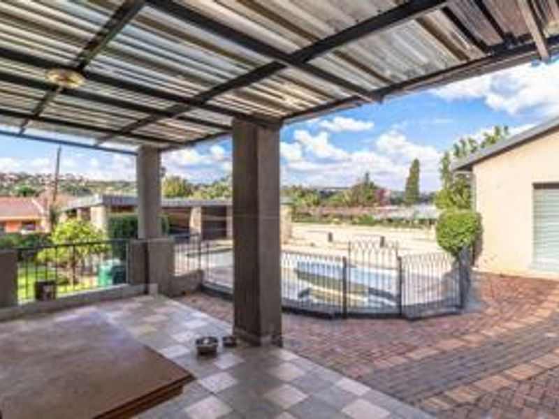 Covered outdoor patio area featuring paved flooring, concrete support pillars, and metal security fencing overlooking the backyard and pool area.