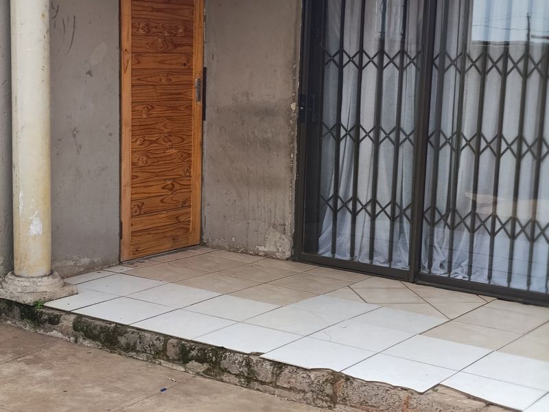 A covered patio area featuring tiled flooring, a sliding glass door with security gate, and a wooden entrance door, requiring maintenance and cosmetic updates.