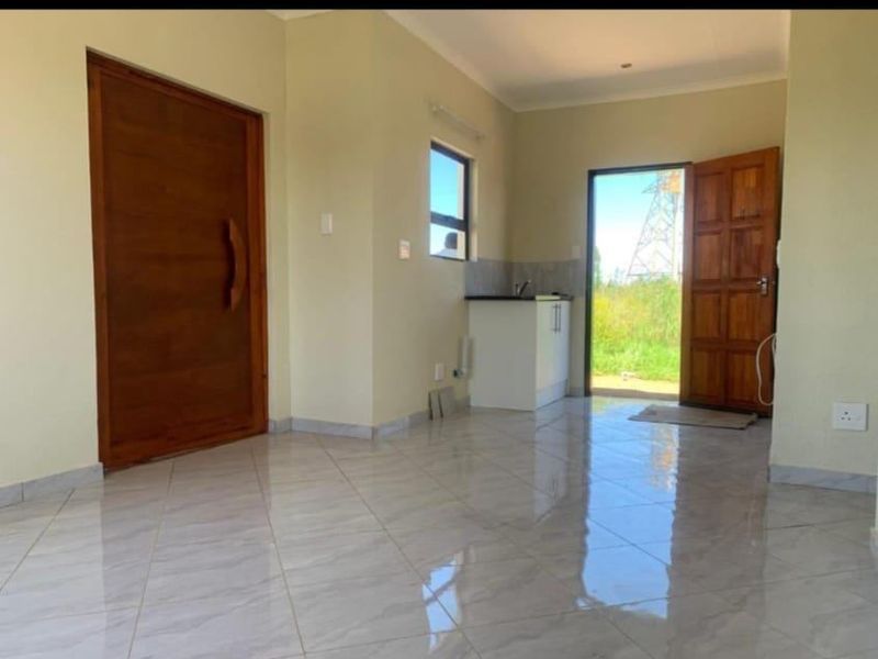 An open-plan living area featuring polished tiled flooring, a wooden entrance door, and a compact kitchen corner with built-in cabinetry and countertop.