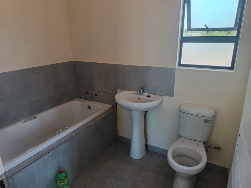 A functional bathroom featuring a bathtub, pedestal basin, and toilet with grey tiled flooring and splashbacks, awaiting final finishing touches.