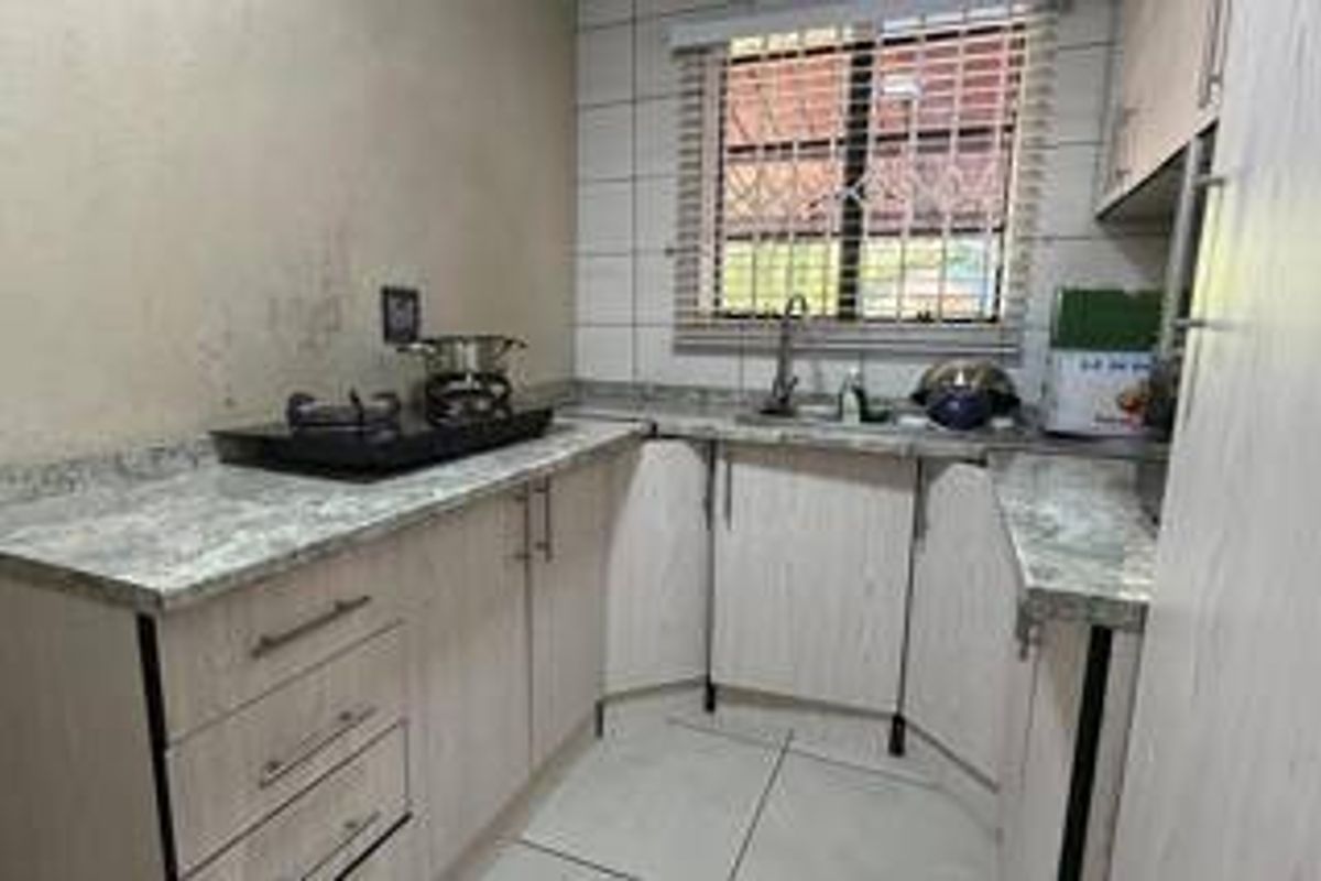 A compact kitchen featuring light-toned laminate cabinetry, stone-look countertops, a stainless steel sink, and tiled splashbacks with security bars on the window.