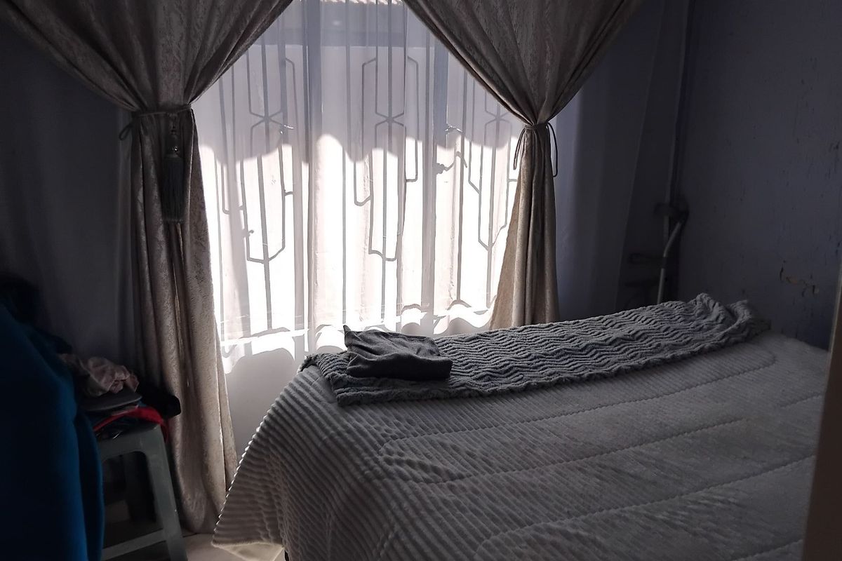A bedroom featuring tiled flooring and security burglar bars on the window, currently showing signs of wear on the wall finishes.