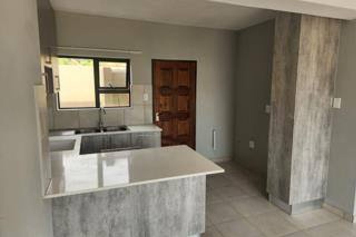 A clean, modern kitchen featuring grey wood-look cabinetry, stone countertops, a double sink, and neutral tiled flooring in an open-plan layout.