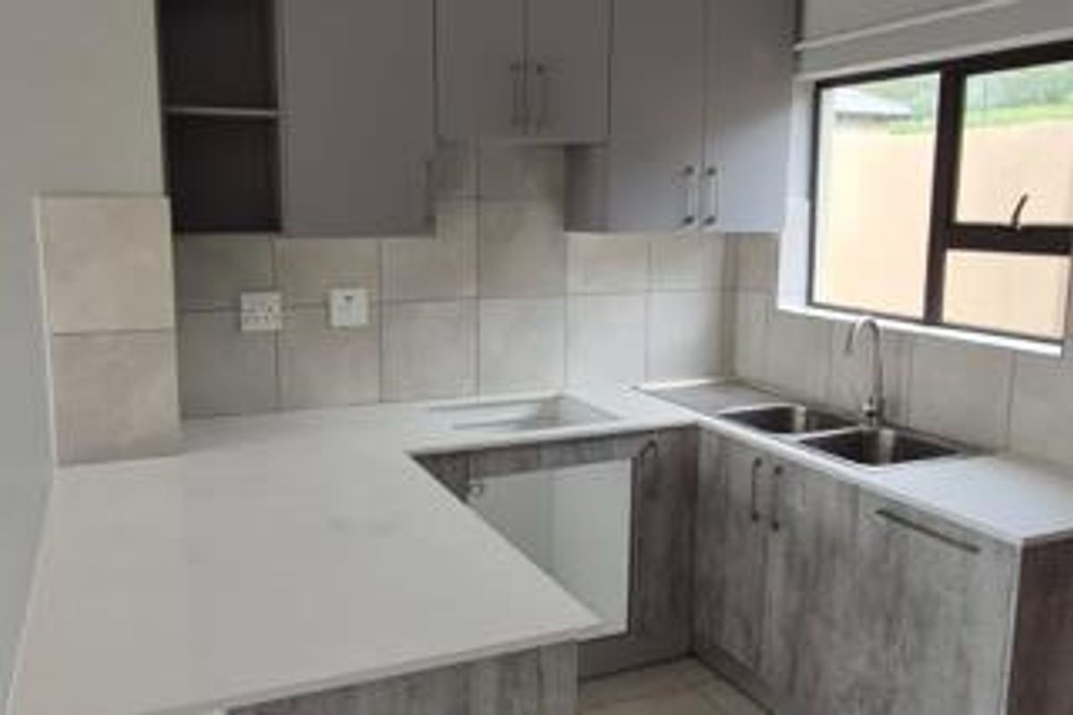 A modern, clean kitchen featuring grey cabinetry, white quartz countertops, a double sink, and a tiled backsplash with ample natural light.