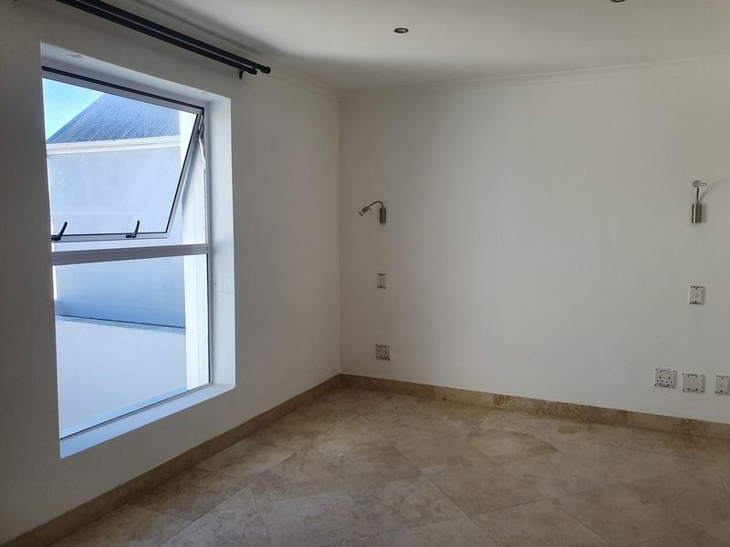An empty bedroom featuring neutral travertine tile flooring, wall-mounted lighting fixtures, and a large window providing natural light.