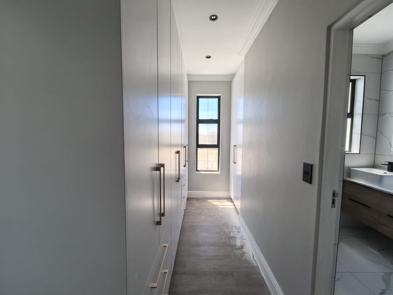 A modern walk-in dressing area featuring extensive floor-to-ceiling built-in cabinetry, recessed lighting, and a window, leading directly into an en-suite bathroom.