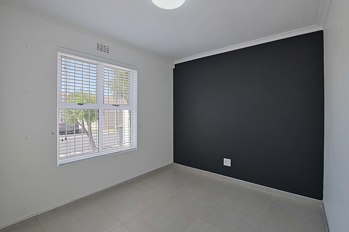 A bright, empty bedroom featuring neutral floor tiles, a contrasting black feature wall, and a window fitted with white venetian blinds.