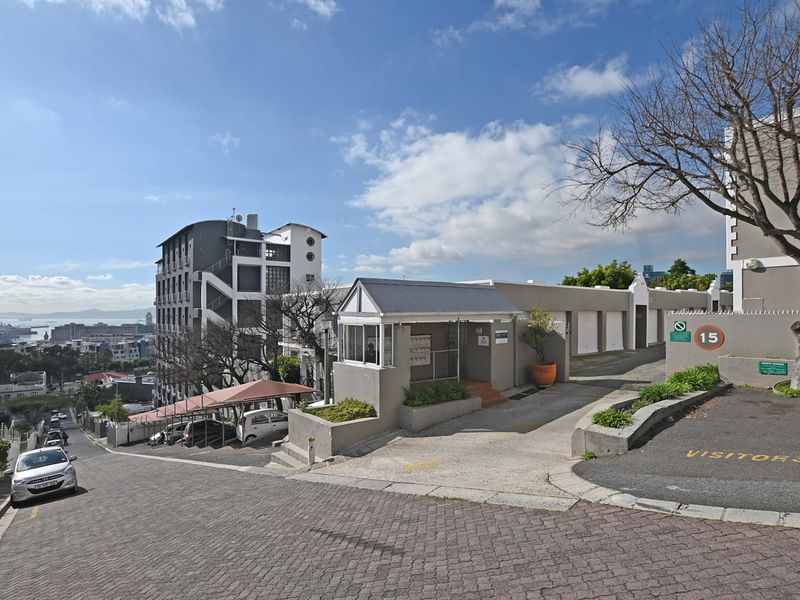Exterior view of the apartment complex entrance, featuring a paved driveway, security gatehouse, and designated visitor parking area.