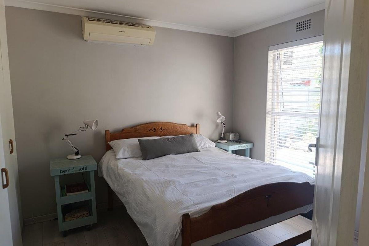 A well-lit bedroom featuring light-toned laminate flooring, a wall-mounted air conditioning unit, and a window with fitted blinds.