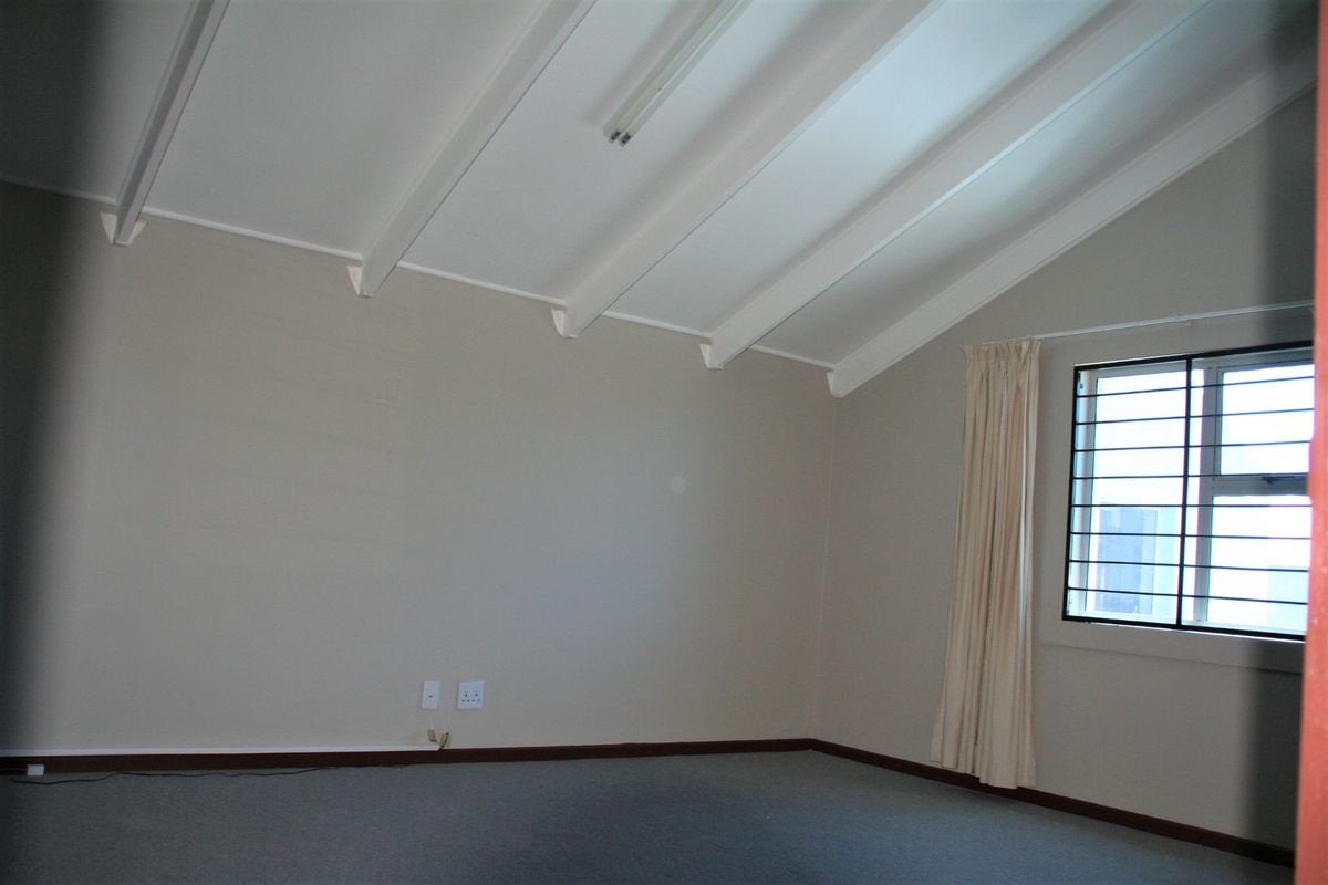 An empty interior room featuring a vaulted ceiling with exposed beams, carpeted flooring, and a window with security bars.