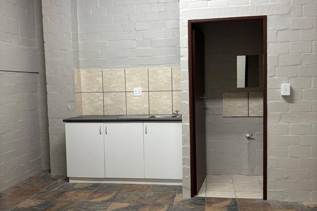 A functional kitchenette area featuring built-in cabinetry, a sink, tiled splashback, and durable floor tiling, adjacent to a small utility or bathroom space.