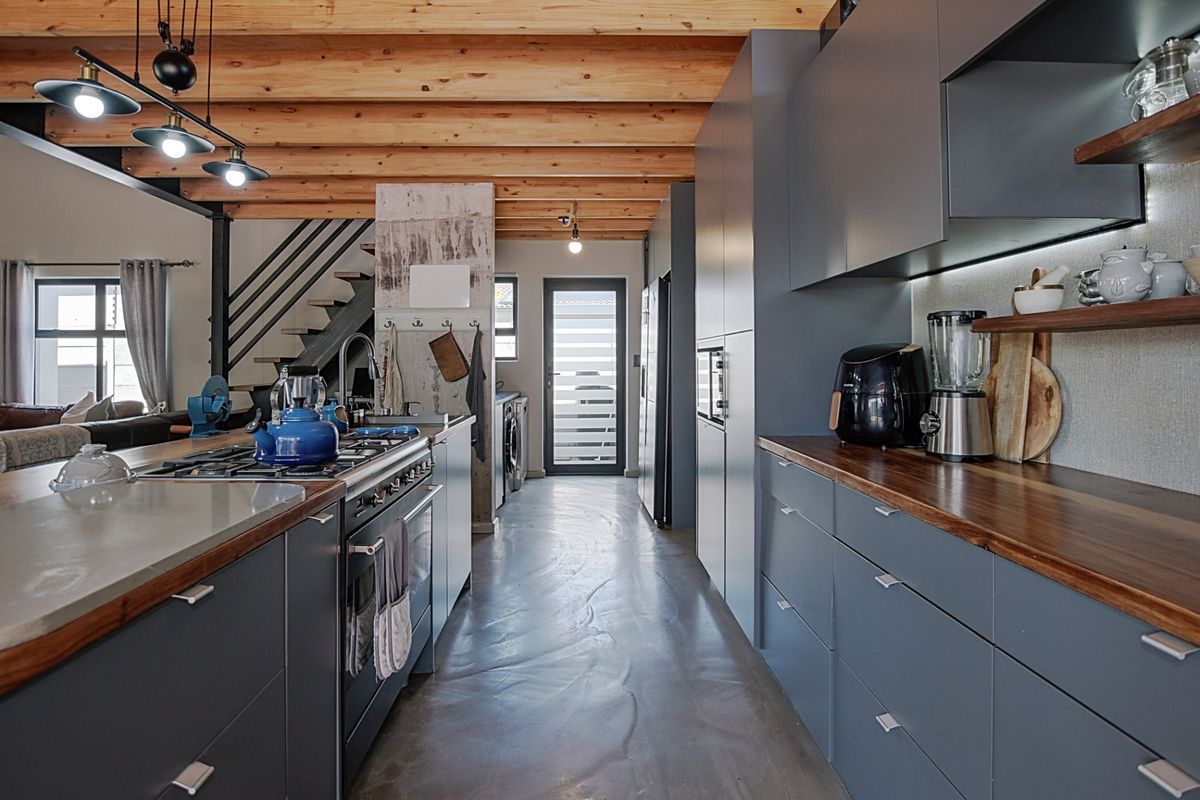 Modern kitchen featuring dark cabinetry, wooden countertops, exposed timber ceiling beams, and polished concrete flooring, seamlessly integrated with an open-plan living area.